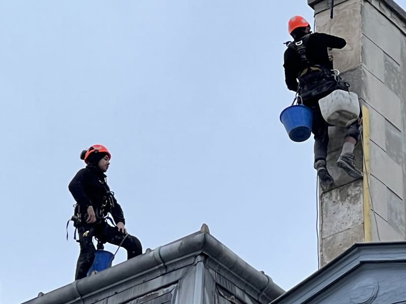 Deux cordistes sécurisés interviennent en hauteur sur une cheminée d’immeuble à Paris, équipés de casques et de seaux de travail.