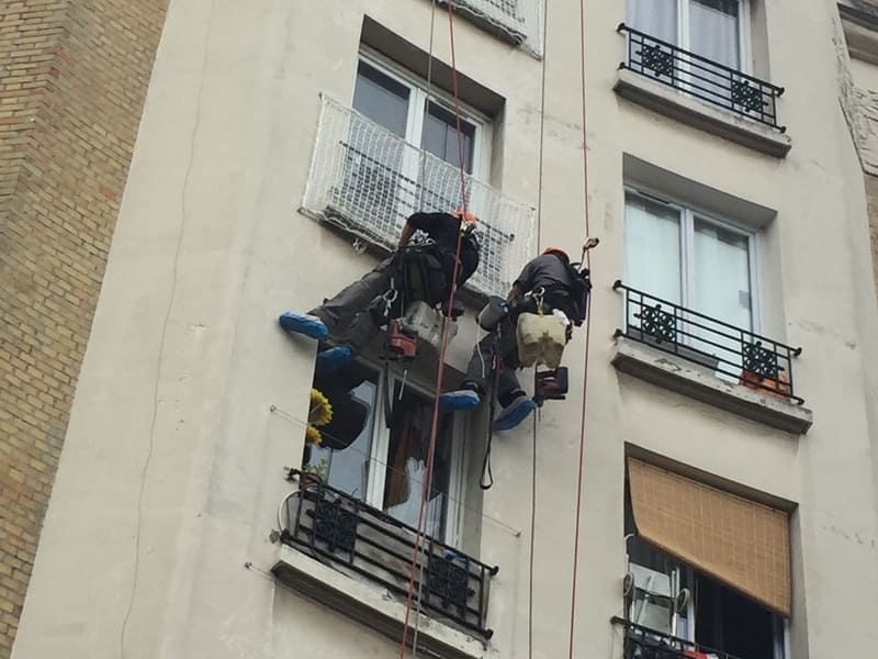 Deux cordistes suspendus en intervention sur la façade d’un immeuble parisien, équipés de harnais, cordes et seaux de travail.
